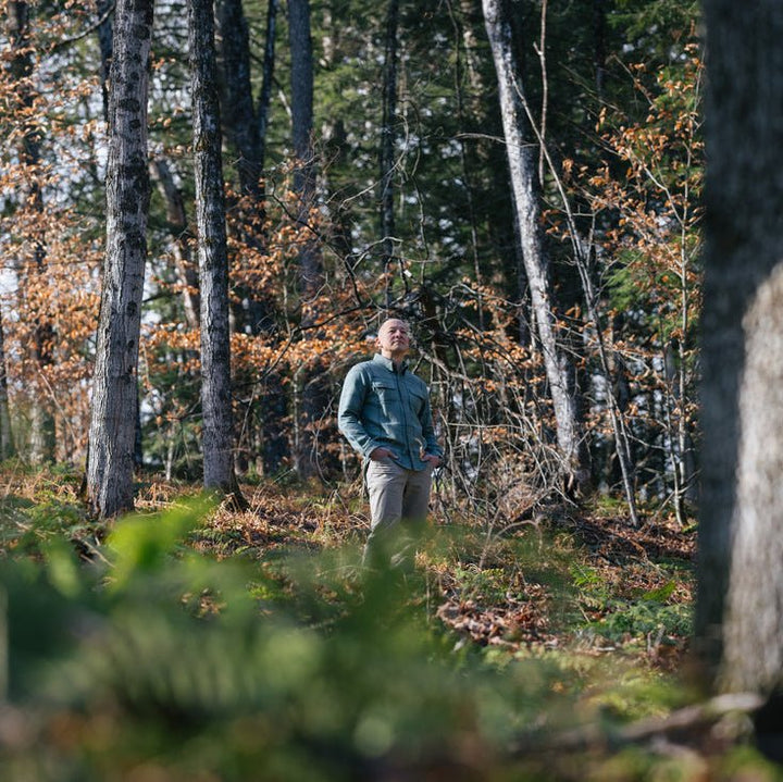 A man wearing a lichen flanneland beige pants stands in a wooded forest clearing with tall trees and fallen autumn leaves.