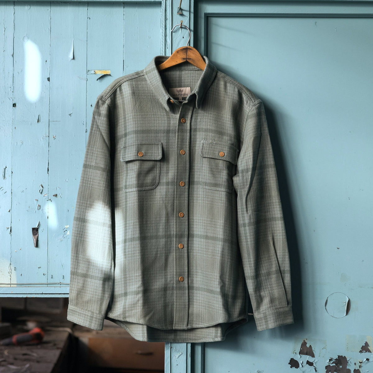 A long-sleeve button-up shirt with a subtle plaid pattern in lichen, hanging on a wooden hanger against a weathered, light blue painted metal door.