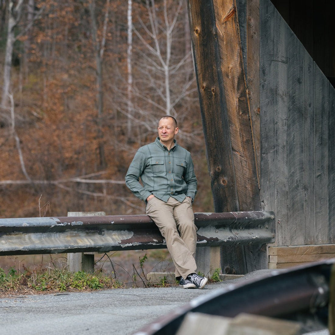 A man wearing a lichen flannel shirt and khaki pants sits on a metal guardrail outdoors next to a large wooden structure, with bare trees in the background.