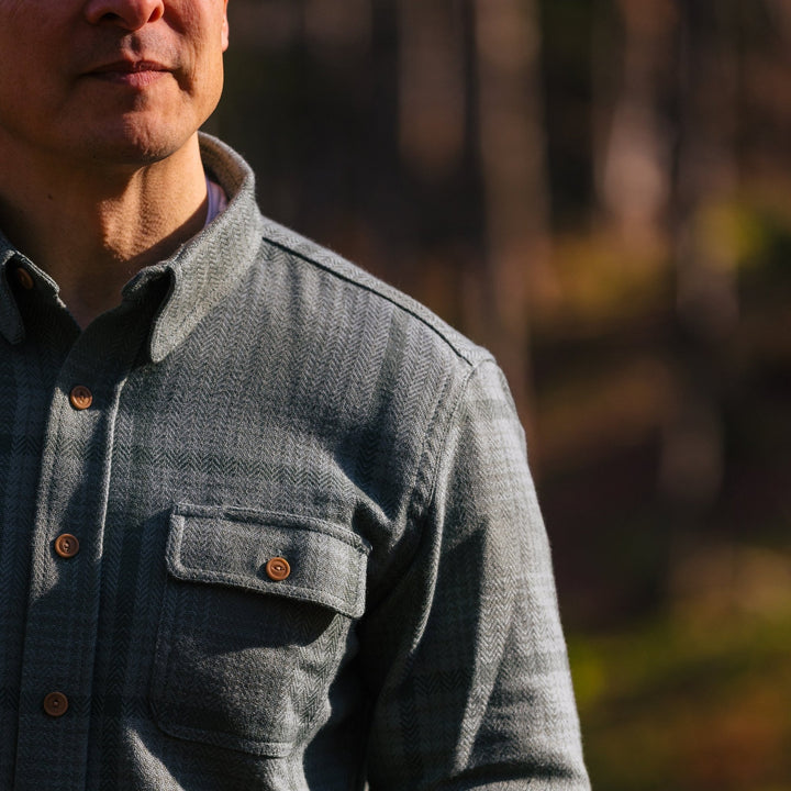 A close-up of a man wearing a textured lichen button-down shirt with brown buttons, set against a blurred outdoor forest background.