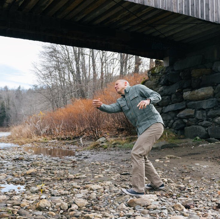 A man in a lichen plaid long-sleeve shirt and tan pants skips rocks on a rocky riverbank under a wooden bridge.