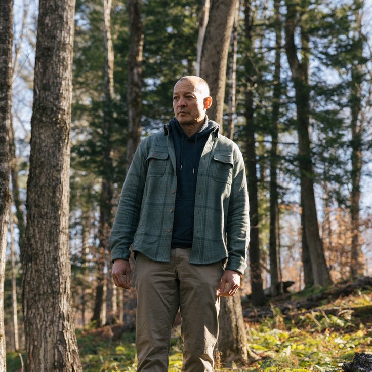 A man standing in a wooded forest wearing a lichen flannel over a dark hoodie and tan pants.