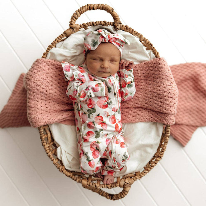 Newborn baby in a Strawberry Organic Snugglesuit Convertible Romper wrapped in a pink blanket inside a wicker basket on a white wooden floor.