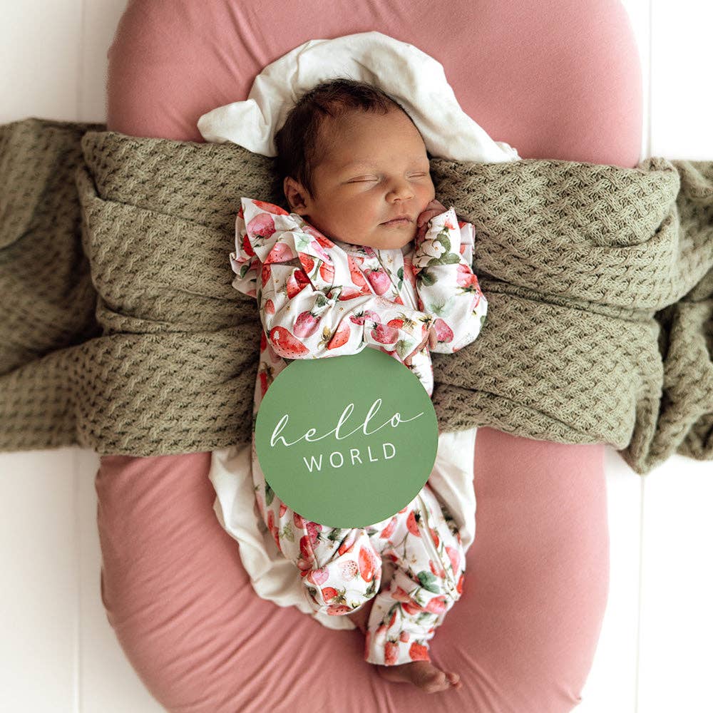 Newborn baby in a Strawberry Organic Snugglesuit Convertible Romper wrapped in a green blanket on a pink cushion on a white wooden floor.
