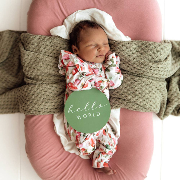 Newborn baby in a Strawberry Organic Snugglesuit Convertible Romper wrapped in a green blanket on a pink cushion on a white wooden floor.