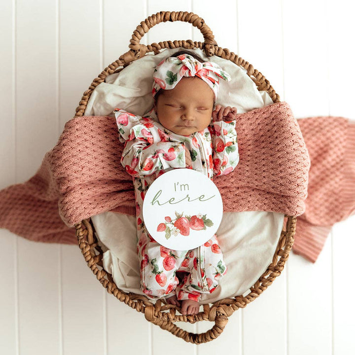 Newborn baby in a Strawberry Organic Snugglesuit Convertible Romper wrapped in a pink blanket inside a wicker basket on a white wooden floor.