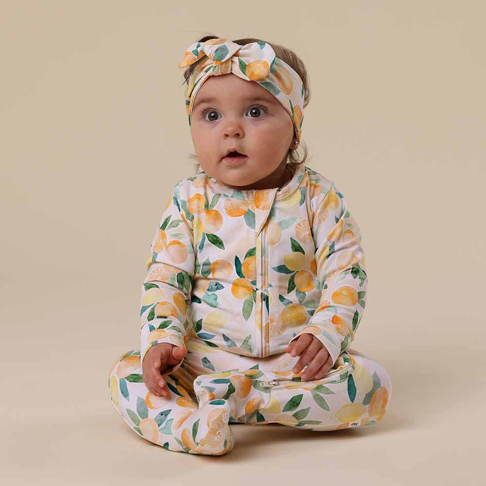 Baby wearing a Sunny Citrus Organic Snuggle Sleepsuit Zip Footie and headband on a beige background