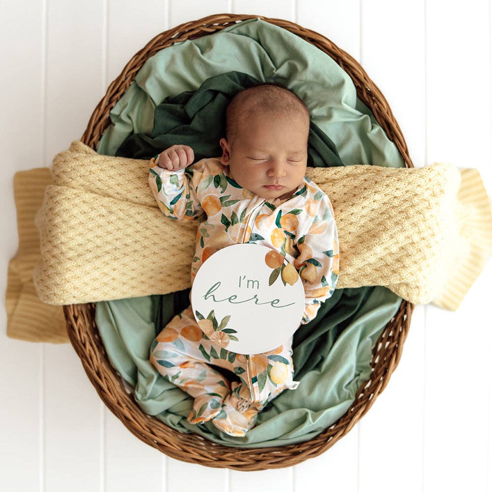 Newborn baby in a Sunny Citrus Organic Snuggle Sleepsuit Zip Footie and hat lying in a woven basket with green and yellow blankets.