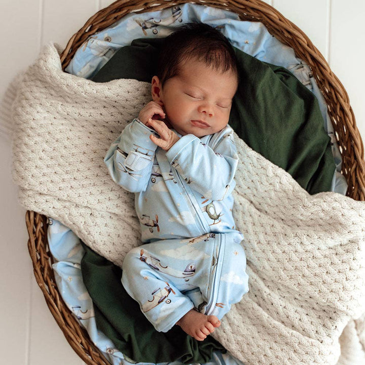 Newborn baby in a Taking Flight Organic Snugglesuit Convertible Romper, sleeping in a woven basket.