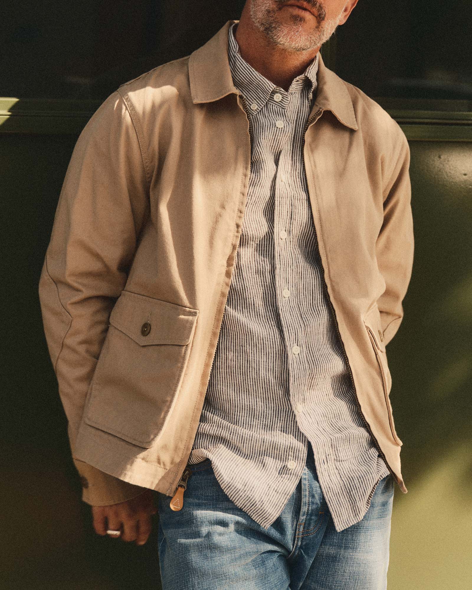 Man wearing Taylor Stitch's Airman Jacket in Organic Khaki Foundation Twill over a striped shirt with a blurred background