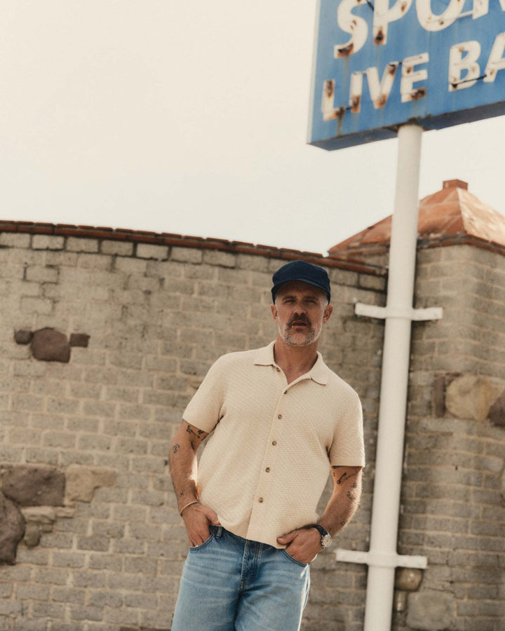 Man wearing The Arlo Sweater Polo
in Oat Texture Knit standing in front of a brick wall with a sign in the background