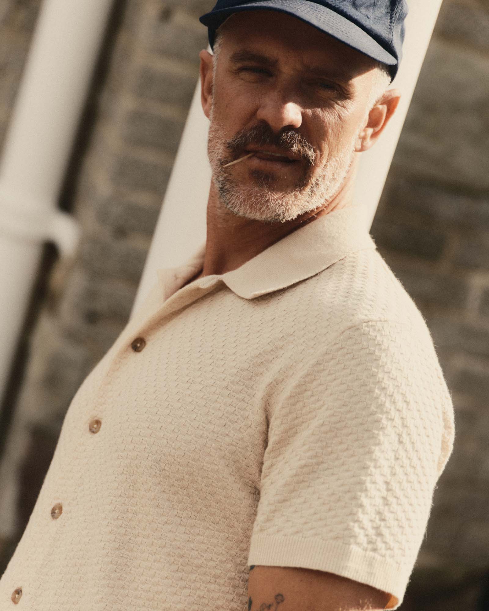 Man wearing The Arlo Sweater Polo
in Oat Texture Knit and cap against a stone wall.
