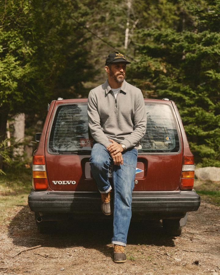 A man wearing a gray quarter-zip pullover, blue jeans, and a black cap sits on the back of a red Volvo SUV in a wooded outdoor setting.