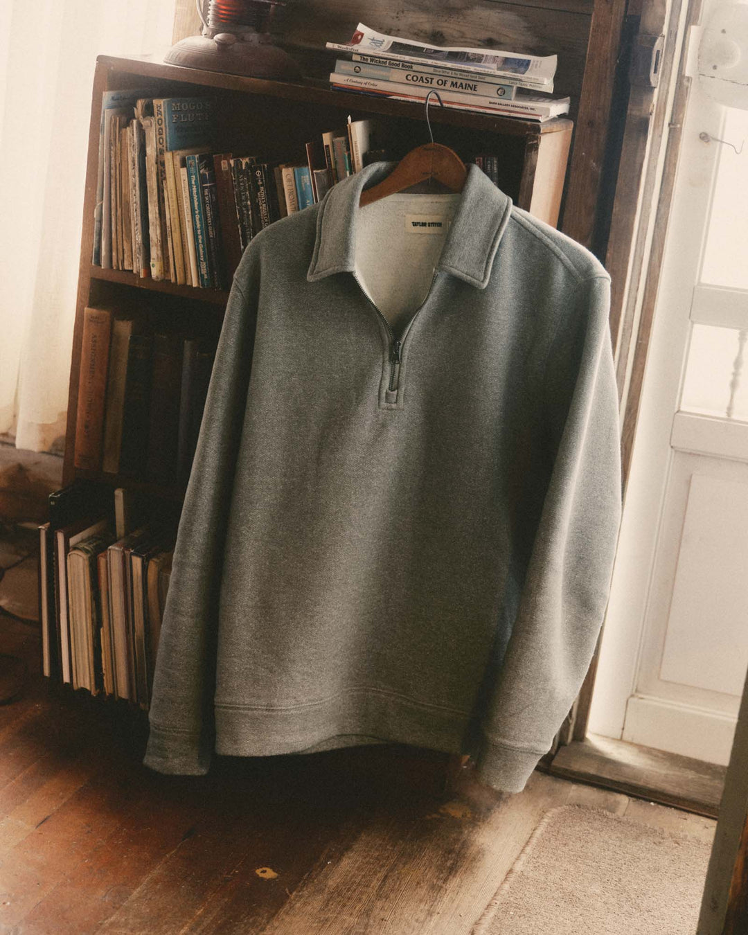 A light gray quarter-zip pullover sweater hanging on a wooden hanger in front of a bookshelf in a rustic indoor setting.