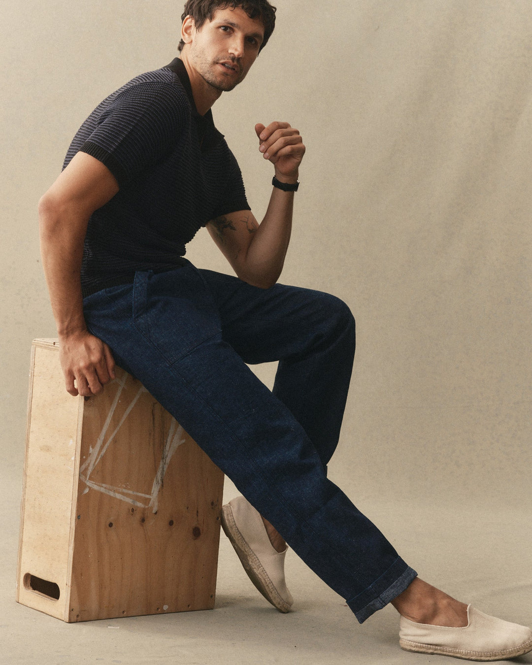 Man sitting on a crate wearing black polo and Taylor Stitch Breakwater pants against a neutral background.
