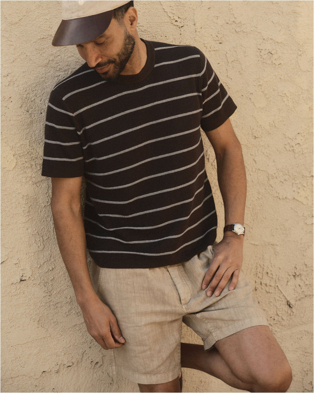 Man wearing The Brody Sweater Tee
in Espresso Stripe and beige shorts against a textured wall.
