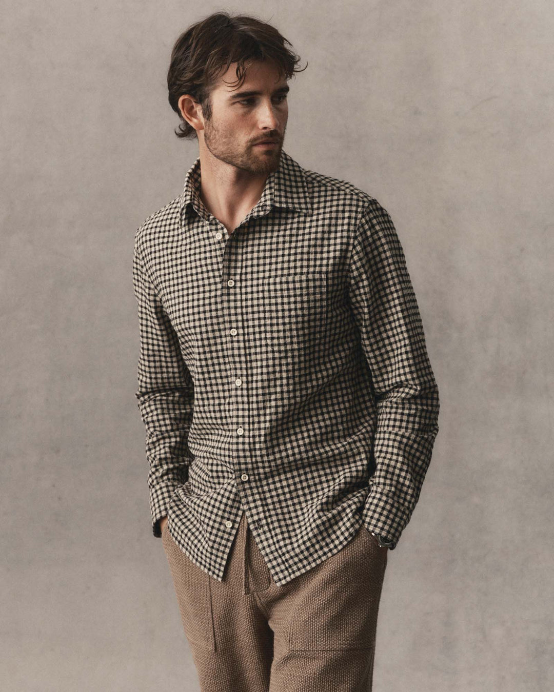 A man wearing The California Shirt
in Breaker Gingham, paired with brown trousers, standing against a textured gray studio background.