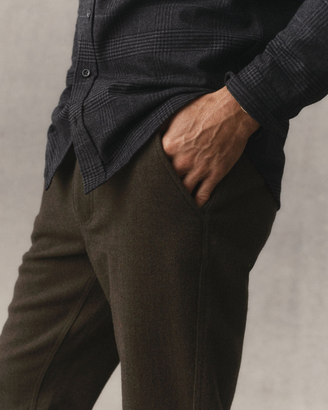 Close up of a man wearing Taylor Stitch Carnegie Pants in Army Wool, with a hand in  the pocket, in front of a grey textured backdrop.