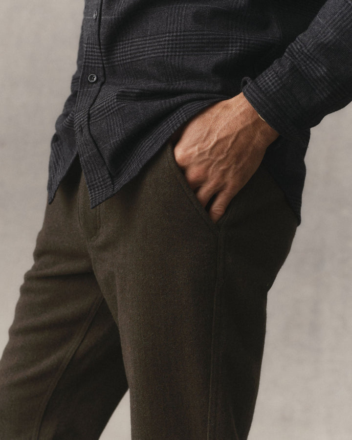 Close up of a man wearing Taylor Stitch Carnegie Pants in Army Wool, with a hand in  the pocket, in front of a grey textured backdrop.