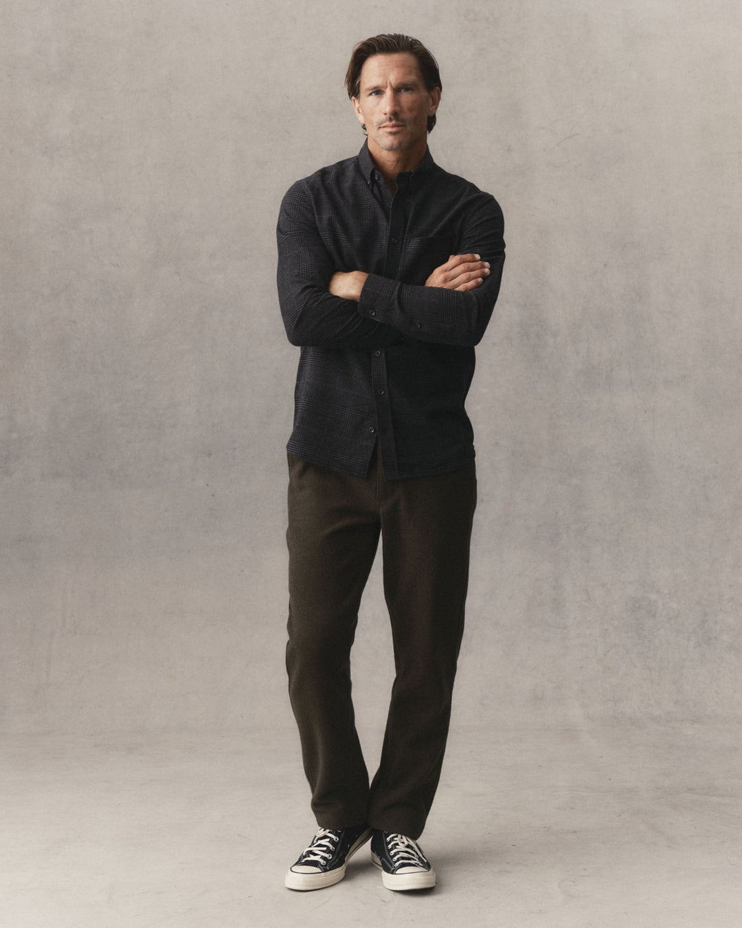 A man wearing Taylor Stitch Carnegie Pants in Army Wool with a dark button up shirt and converse shoes in front of a grey textured backdrop.