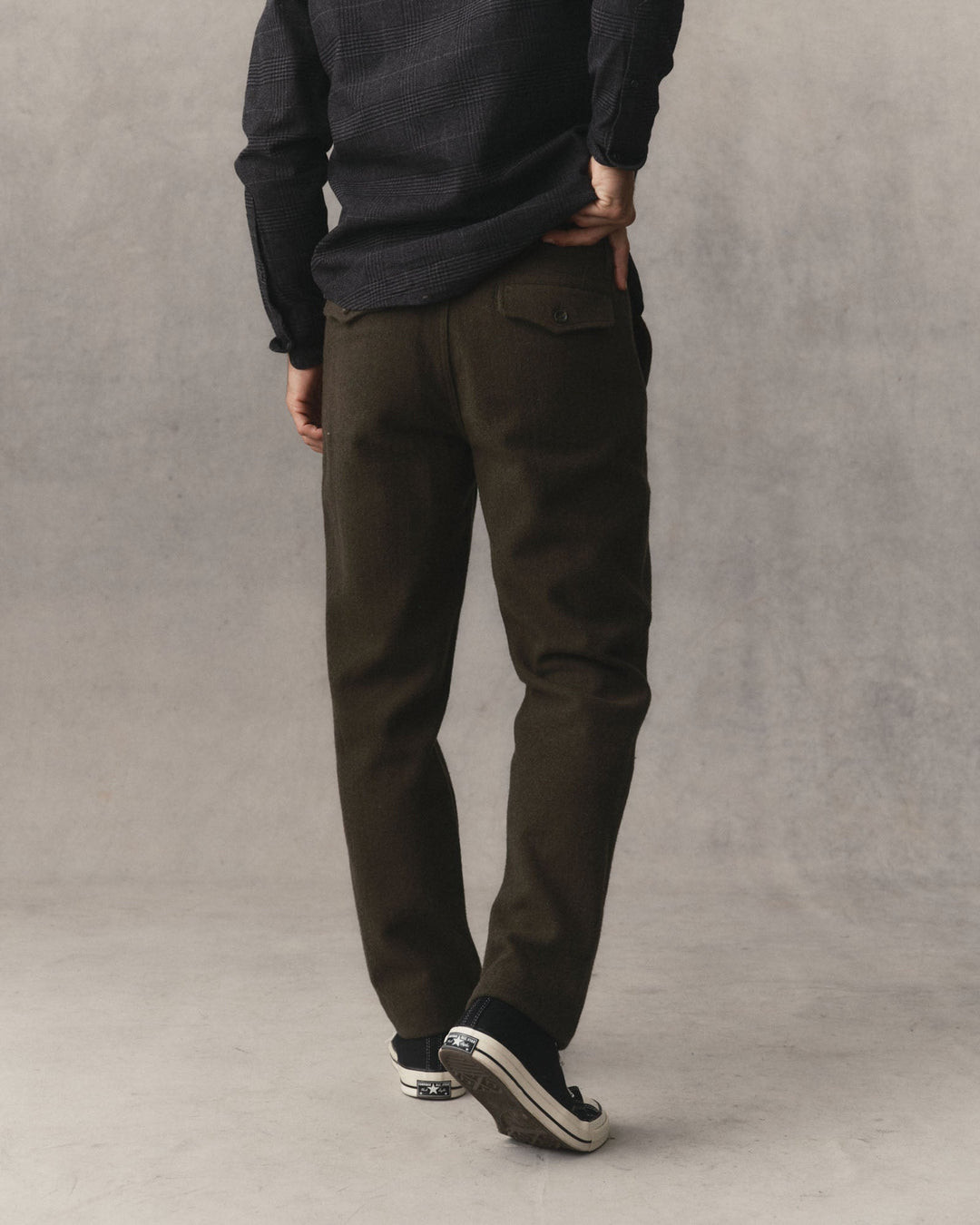 A man shown from behind wearing Taylor Stitch Carnegie Pants in Army Wool with a dark button up shirt and converse shoes in front of a grey textured backdrop.