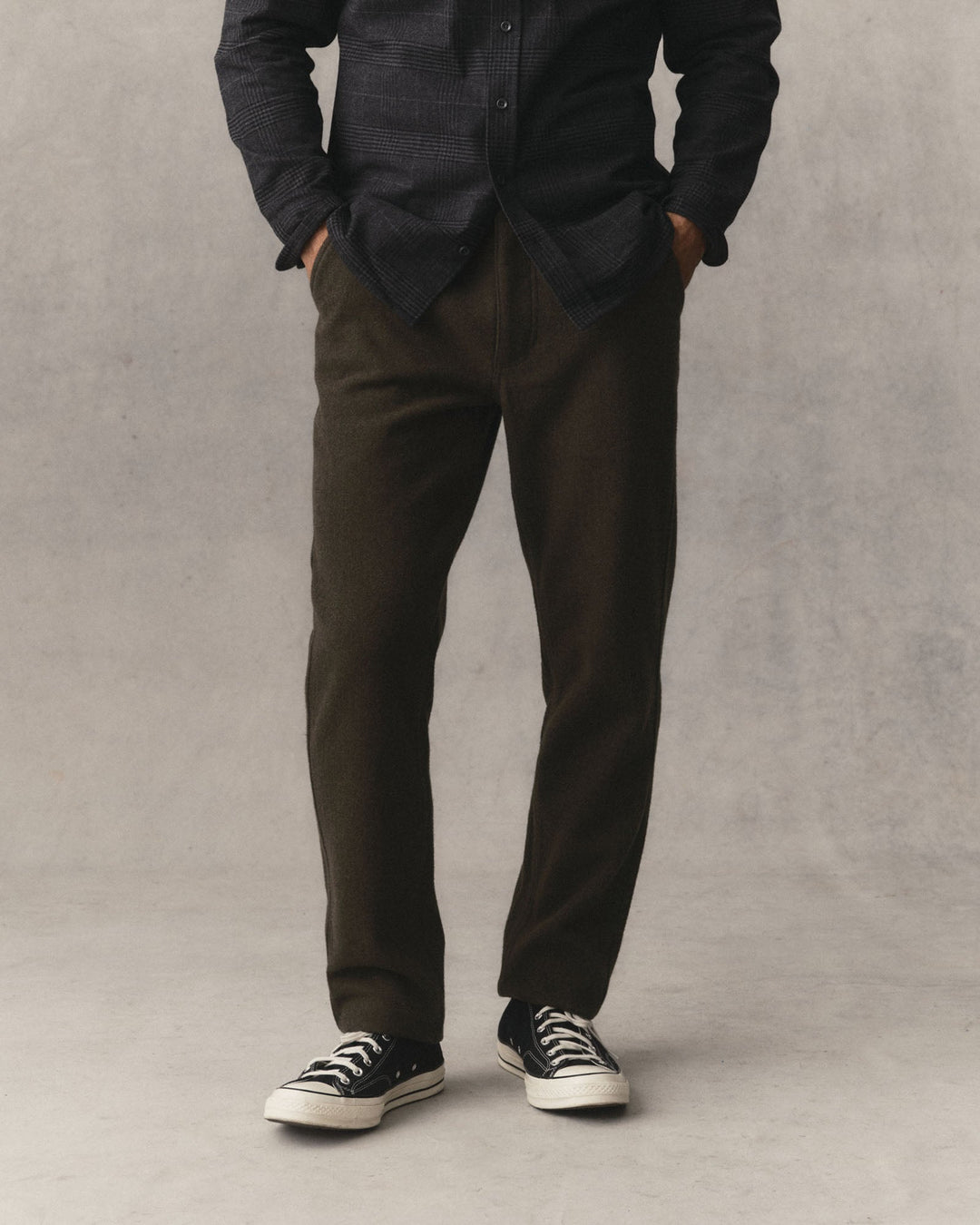 Man wearing Taylor Stitch Carnegie Pant in Army Wool and converse shoes on a grey textured background.