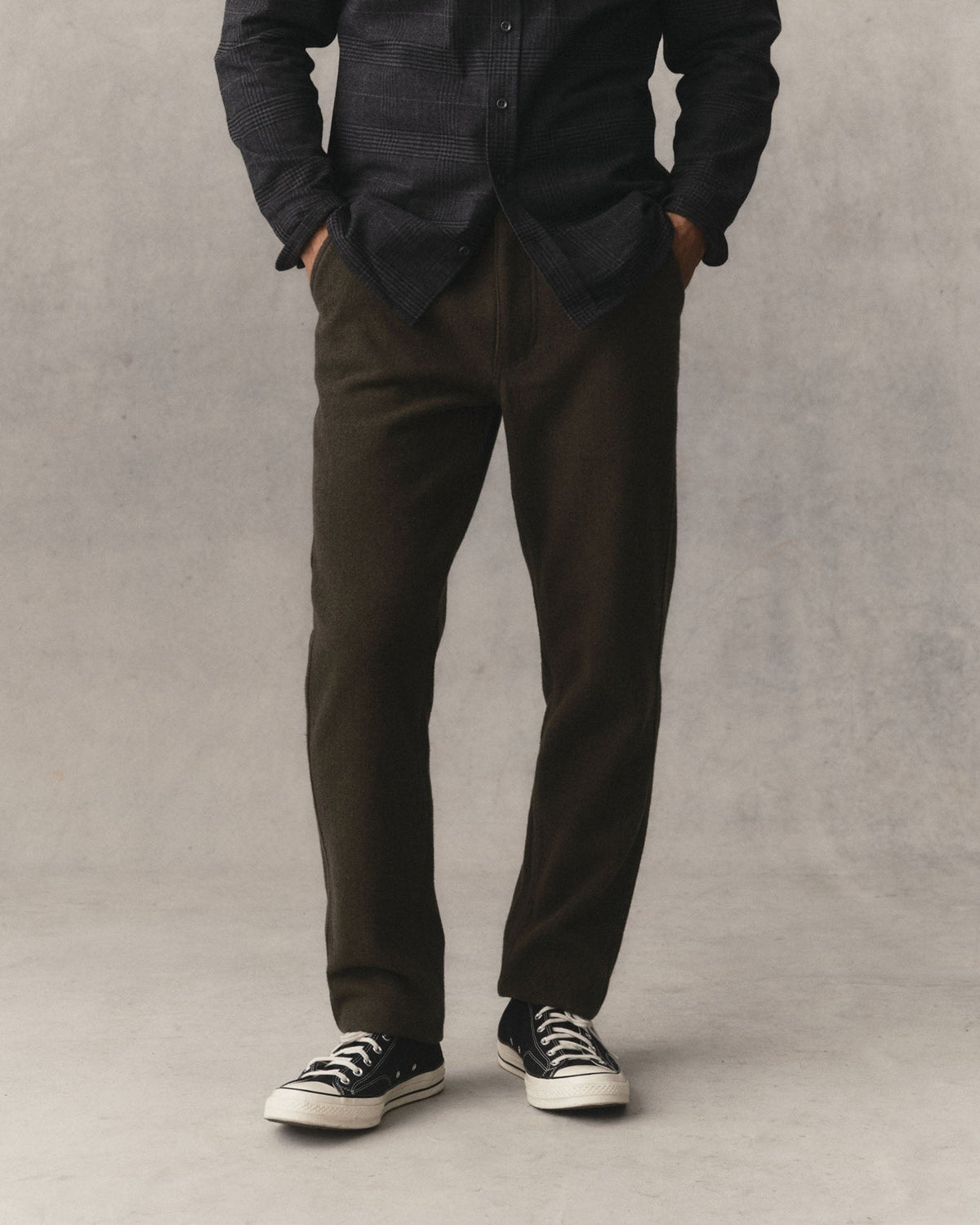 Man wearing Taylor Stitch Carnegie Pant in Army Wool and converse shoes on a grey textured background.