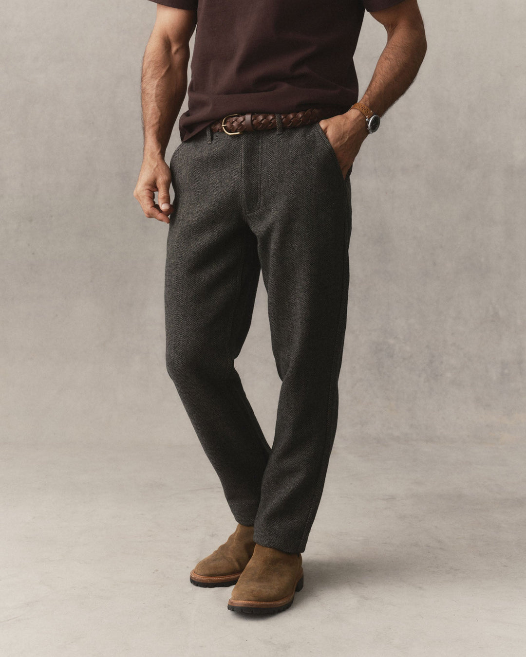A man wearing The Carnegie Pant
in Walnut Herringbone Wool, styled with a brown belt, a dark brown t-shirt, and brown suede boots, standing against a textured gray studio backdrop.