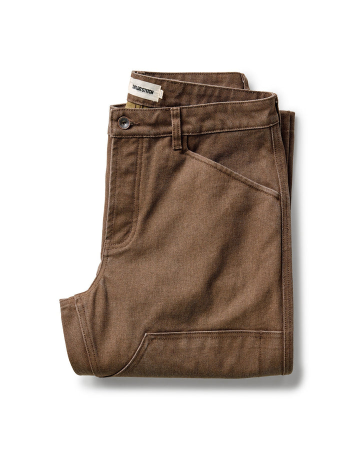 A pair of folded Taylor Stitch Chore Pants in Aged Penny Chipped Canvas with a button closure and belt loops, displayed on a clean white background.