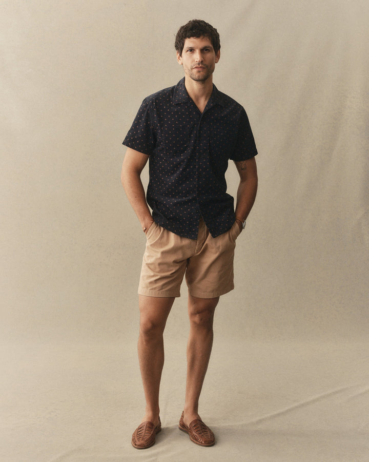 Man wearing a dark patterned shirt and beige shorts against a plain background