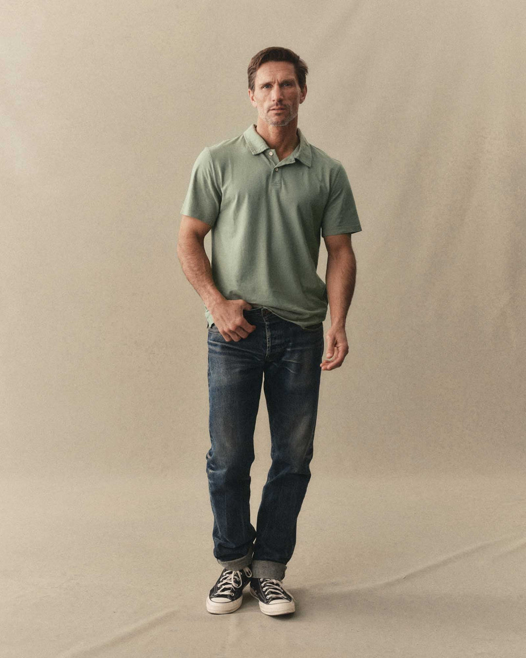 Man wearing a The Cotton Hemp Polo in Sage and blue jeans holding a green bottle against a beige background