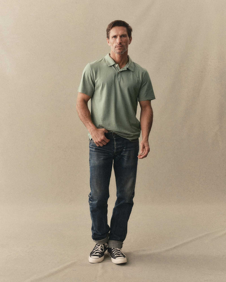 Man wearing a The Cotton Hemp Polo in Sage and blue jeans holding a green bottle against a beige background