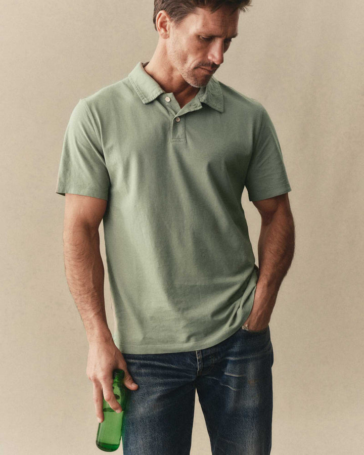 Man wearing a The Cotton Hemp Polo in Sage and blue jeans holding a green bottle against a beige background