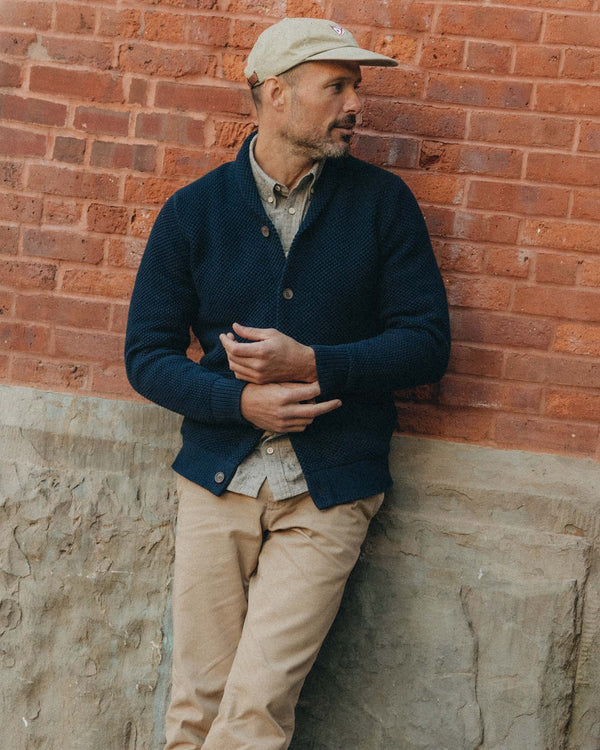 Man wearing Taylor Stitch The Crawford Sweater in Navy and beige cap leaning against a brick wall.