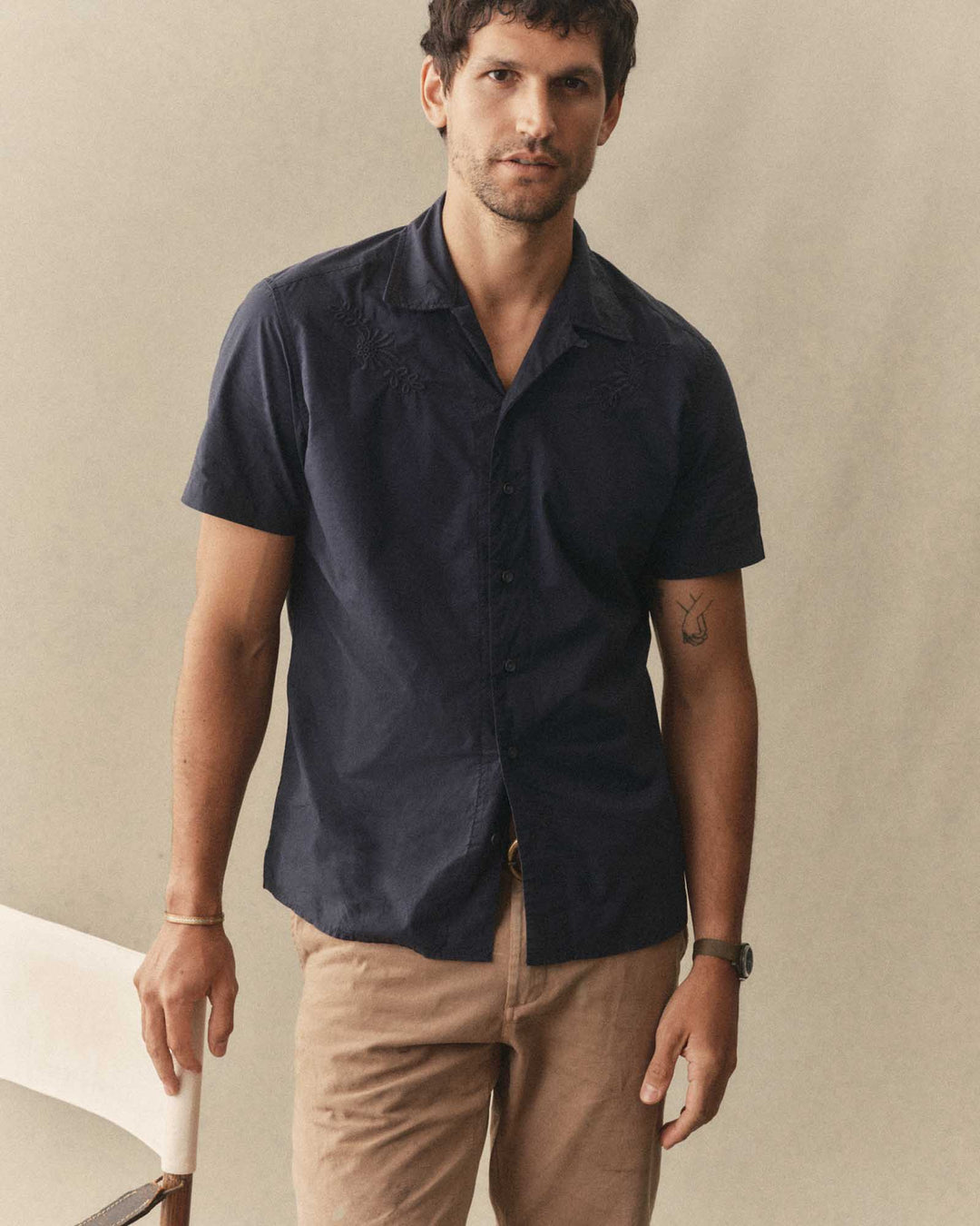 Man wearing The Davis Shirt in Navy Floral Embroidery and beige pants against a beige background