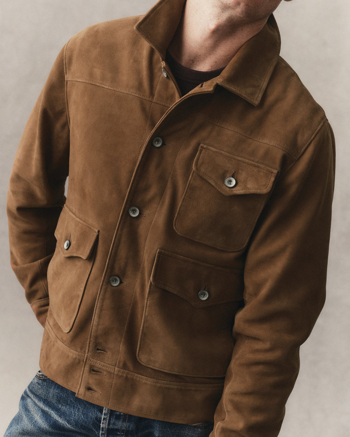 A man wearing Tyalor Stitch's Drover suede Jacket with a collar, snap-button front, and four front flap pockets, paired with blue jeans against a neutral gray background.