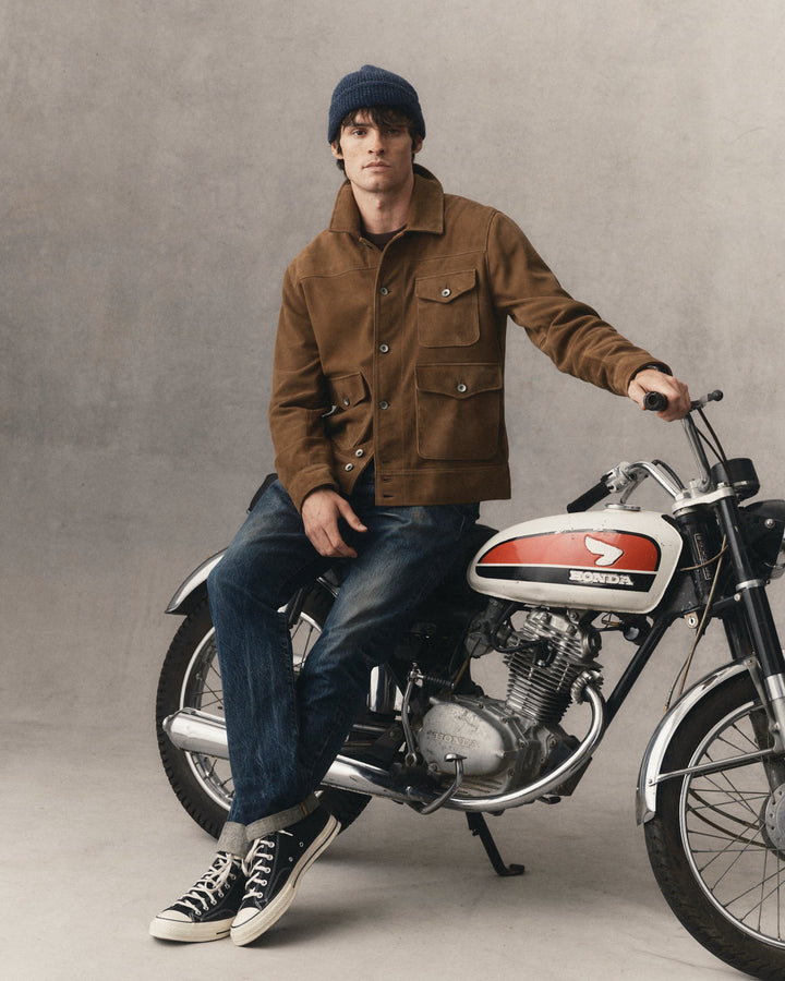 A man wearing a brown jacket, blue jeans, a navy beanie, and black canvas sneakers sits on a vintage Honda motorcycle against a textured gray studio backdrop.
