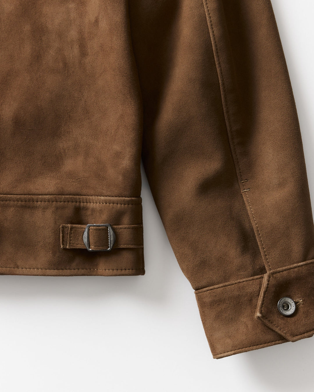 Close-up of a brown suede jacket sleeve showing a buttoned cuff and a matching strap with a metal buckle on the lower cuff, photographed on a light neutral background.