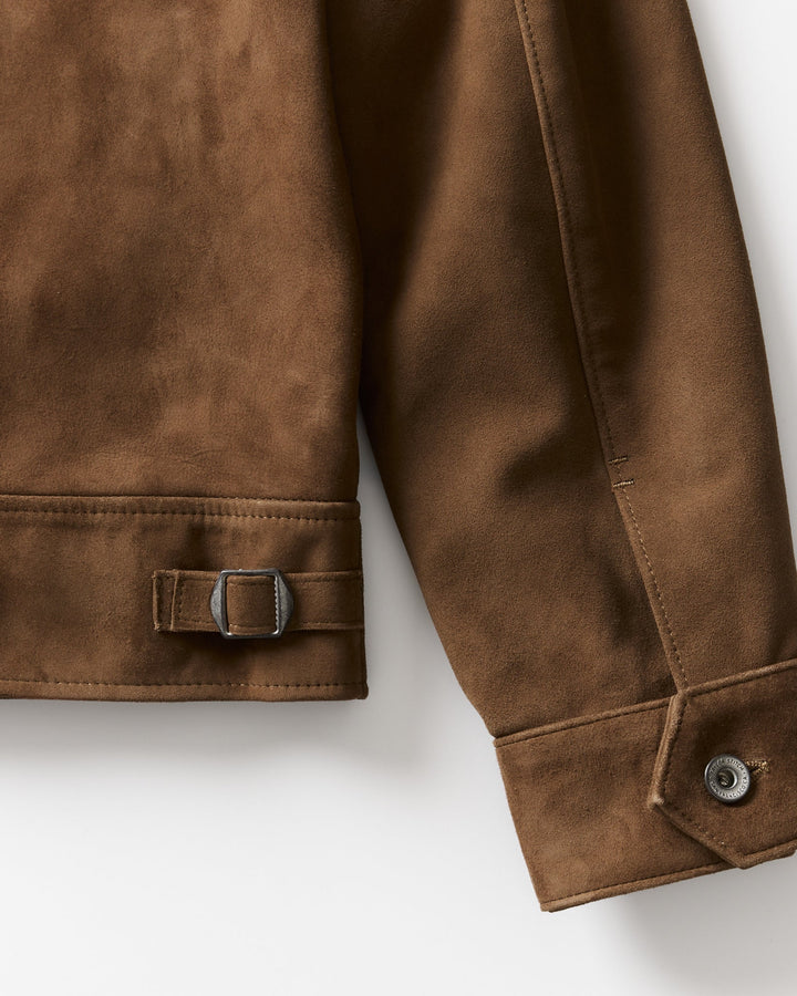 Close-up of a brown suede jacket sleeve showing a buttoned cuff and a matching strap with a metal buckle on the lower cuff, photographed on a light neutral background.