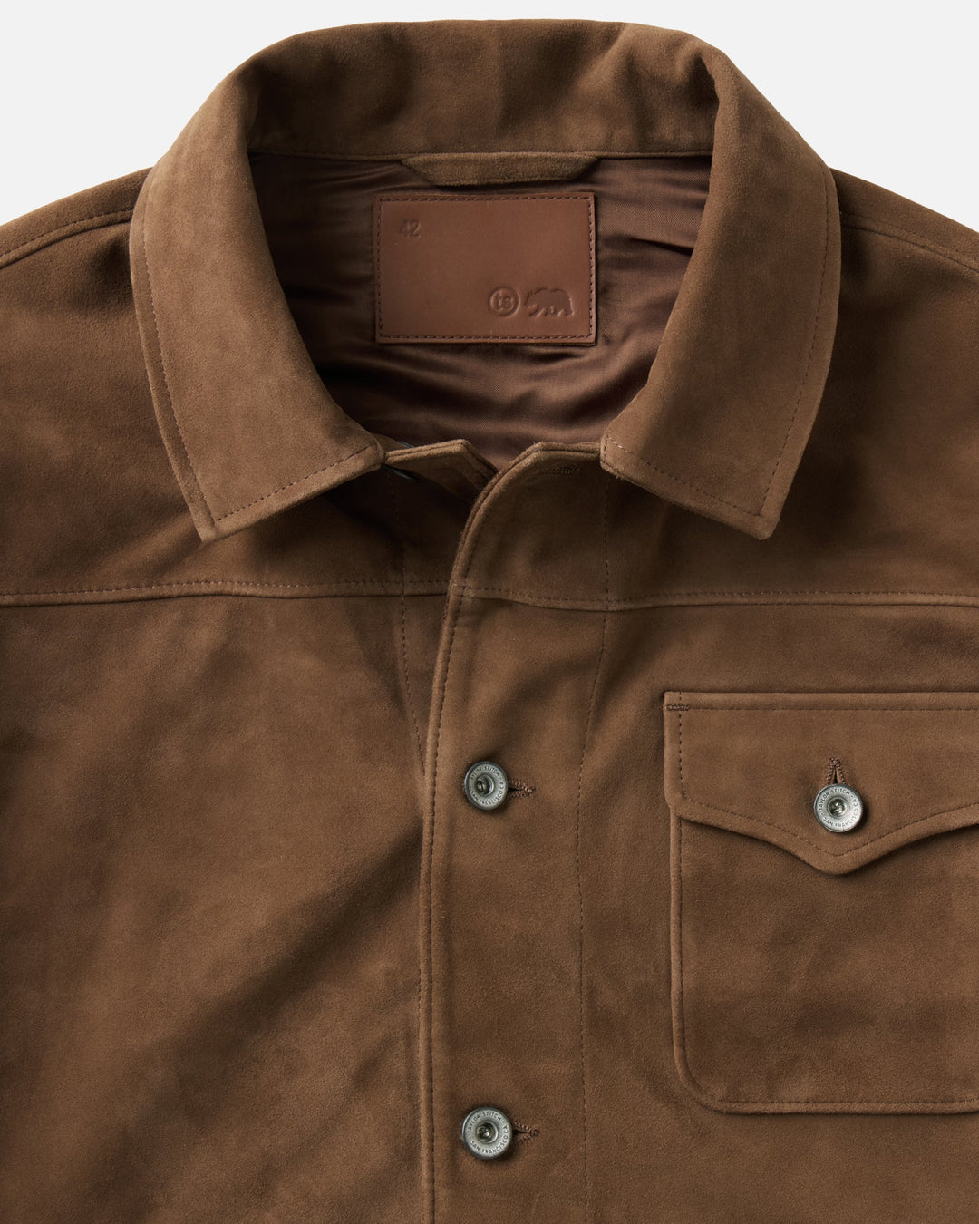 Close-up of a brown suede jacket with a pointed collar, chest pocket, and visible Taylor Stitch brand label inside the collar.