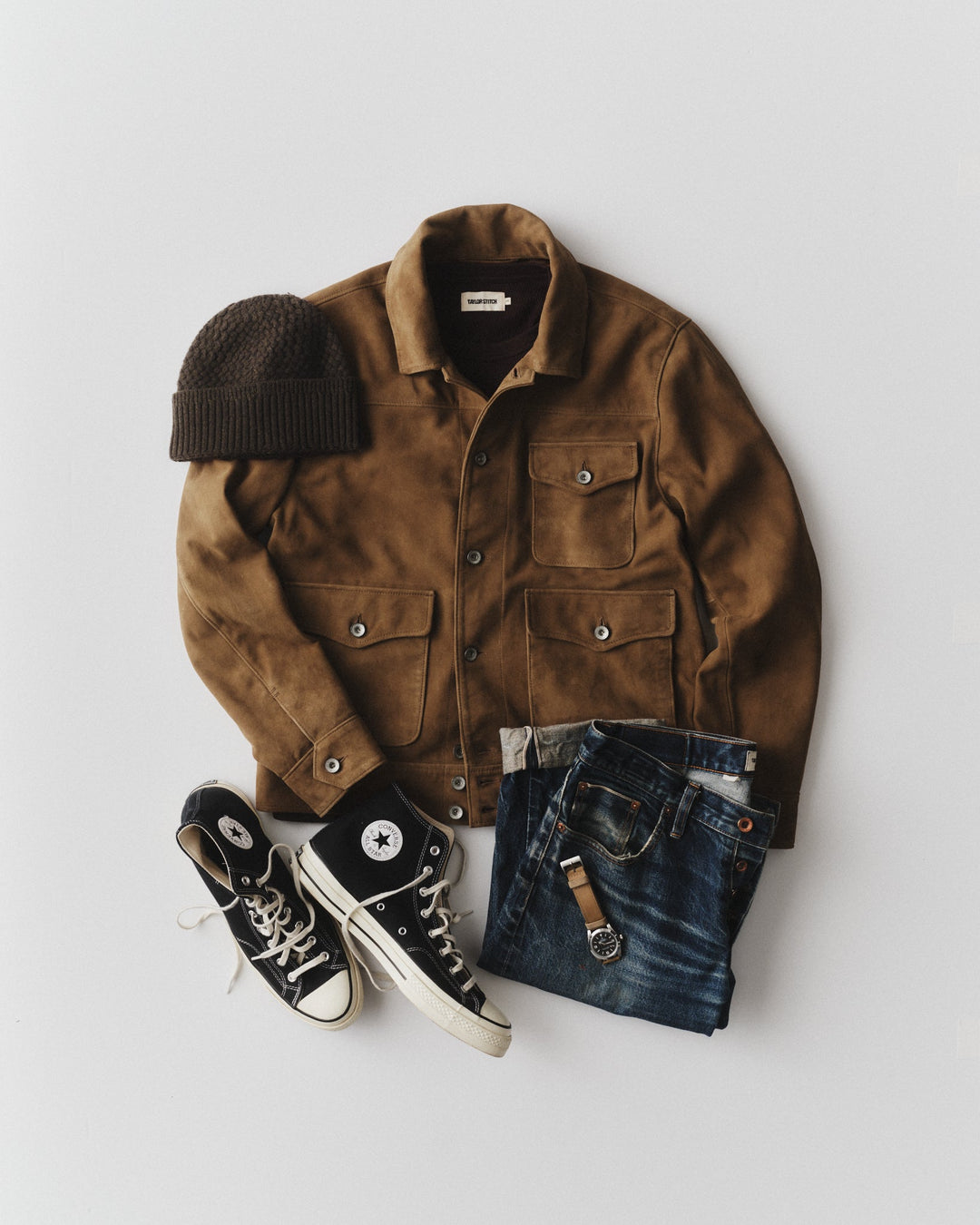A styled flat-lay outfit on a light gray background featuring a brown corduroy jacket, a dark brown knit beanie, black Converse high-top sneakers, and folded blue jeans with a brown leather belt.