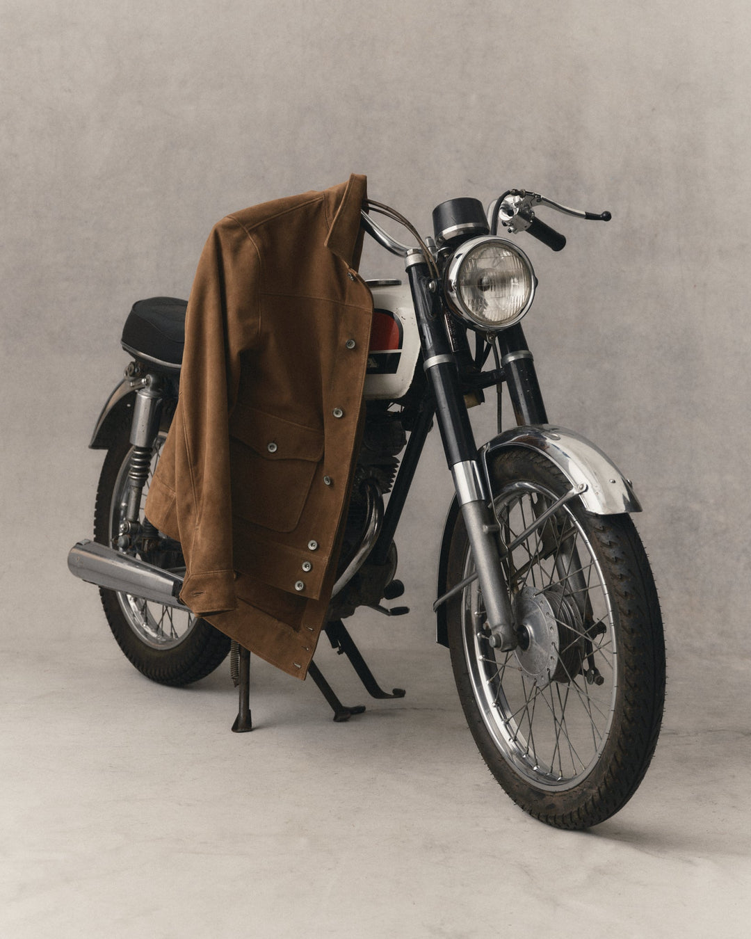 A vintage-style motorcycle photographed in a studio setting with a brown jacket draped over the left side of the frame.