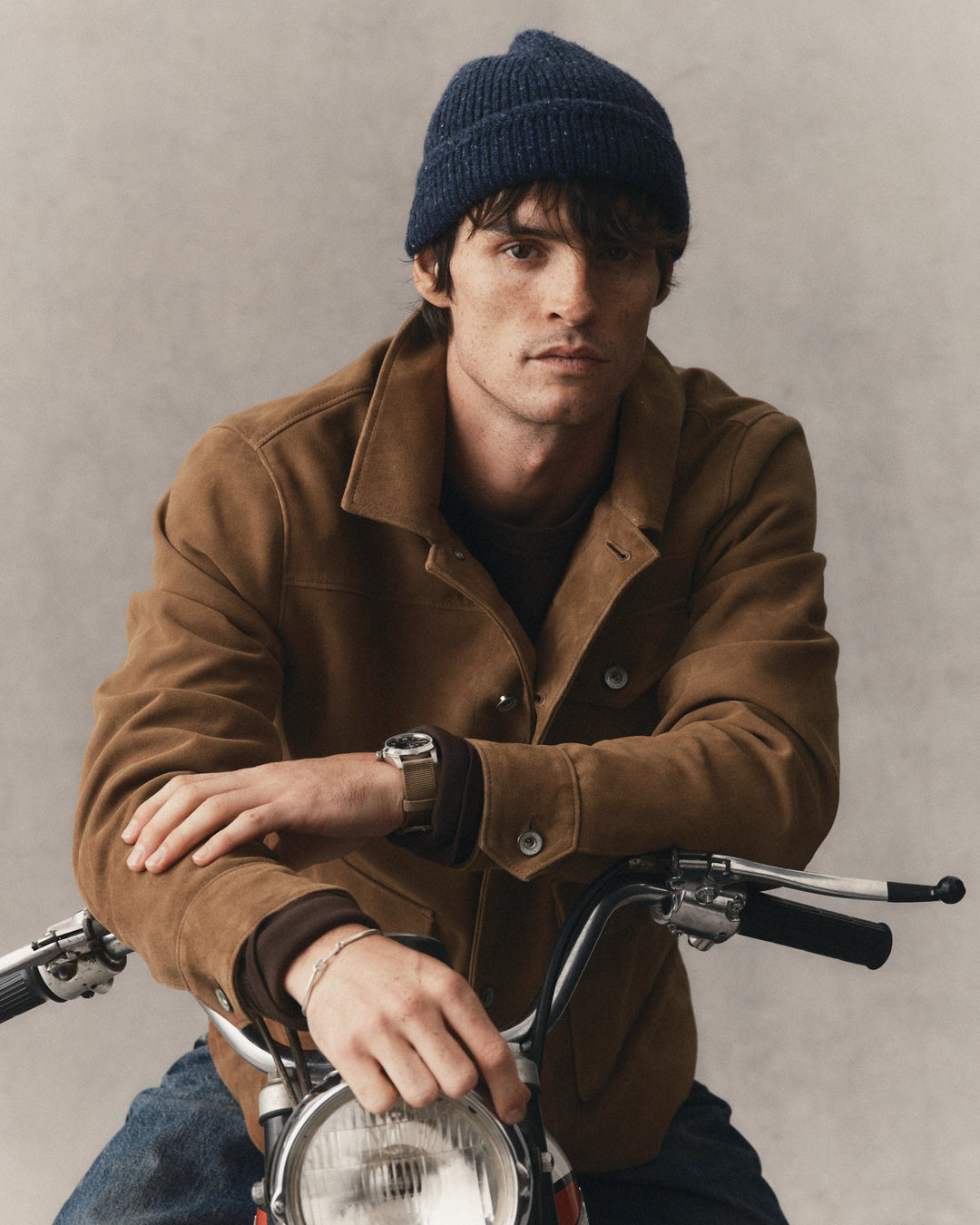A man wearing a dark navy knit beanie, a brown suede jacket, and a watch, leaning on a vintage motorcycle against a textured gray background.
