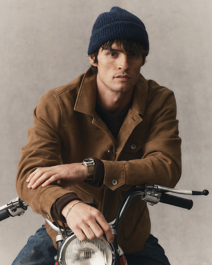 A man wearing a dark navy knit beanie, a brown suede jacket, and a watch, leaning on a vintage motorcycle against a textured gray background.