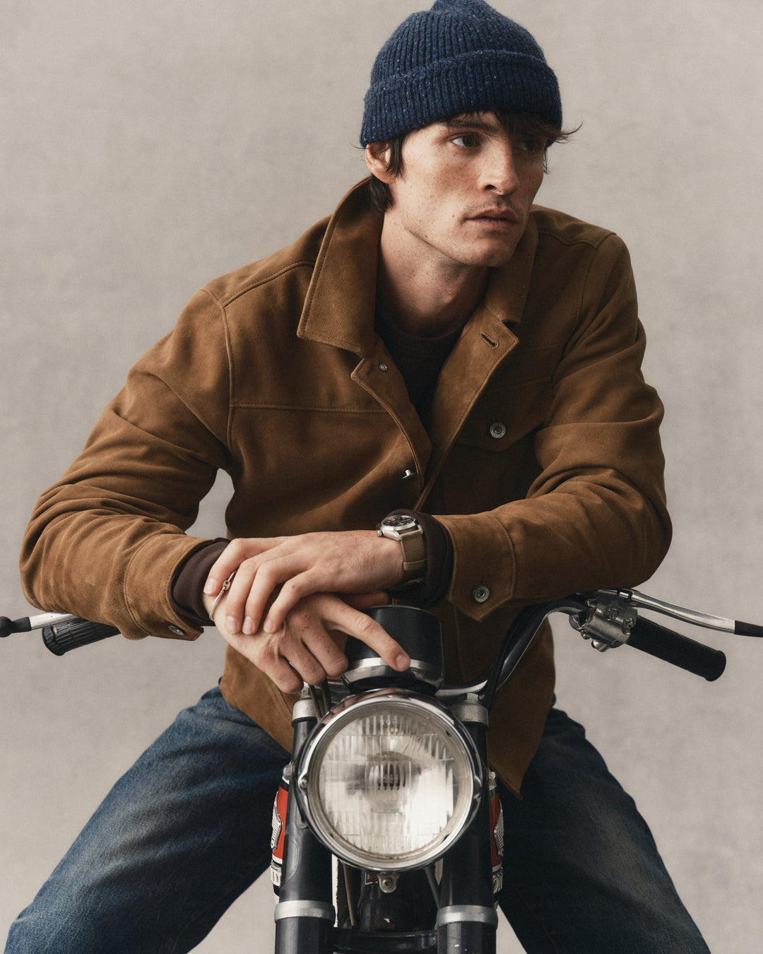 A man wearing a brown suede jacket, blue jeans, and a navy blue beanie sits on a motorcycle against a textured gray background.