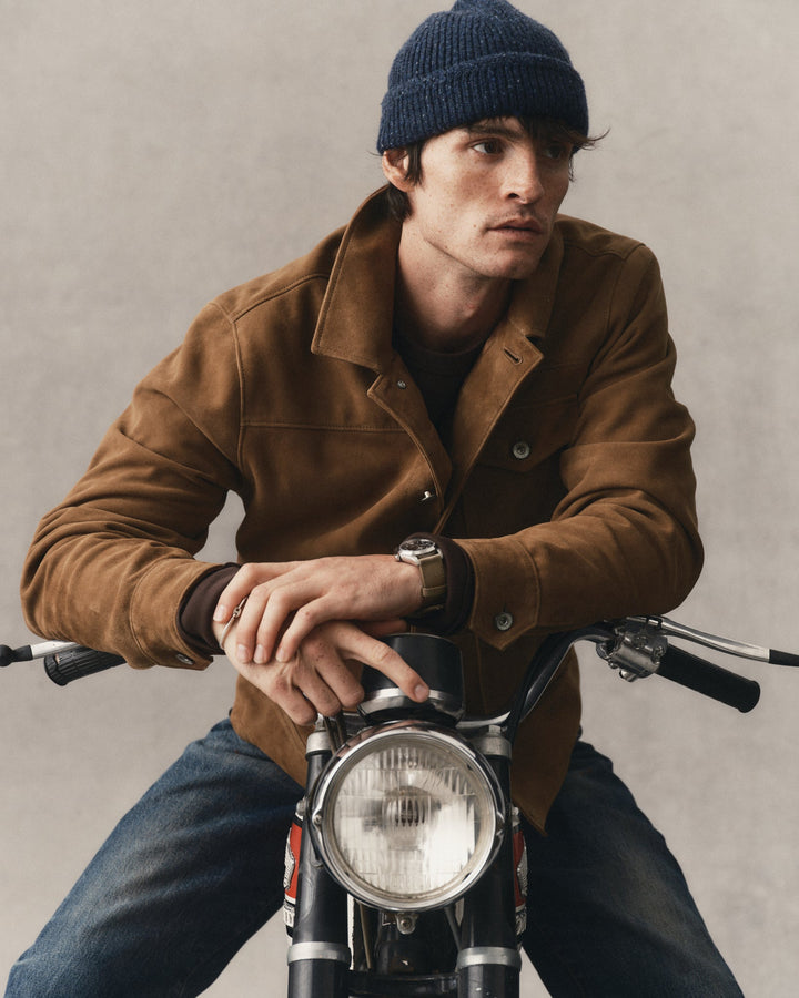 A man wearing a brown suede jacket, blue jeans, and a navy blue beanie sits on a motorcycle against a textured gray background.