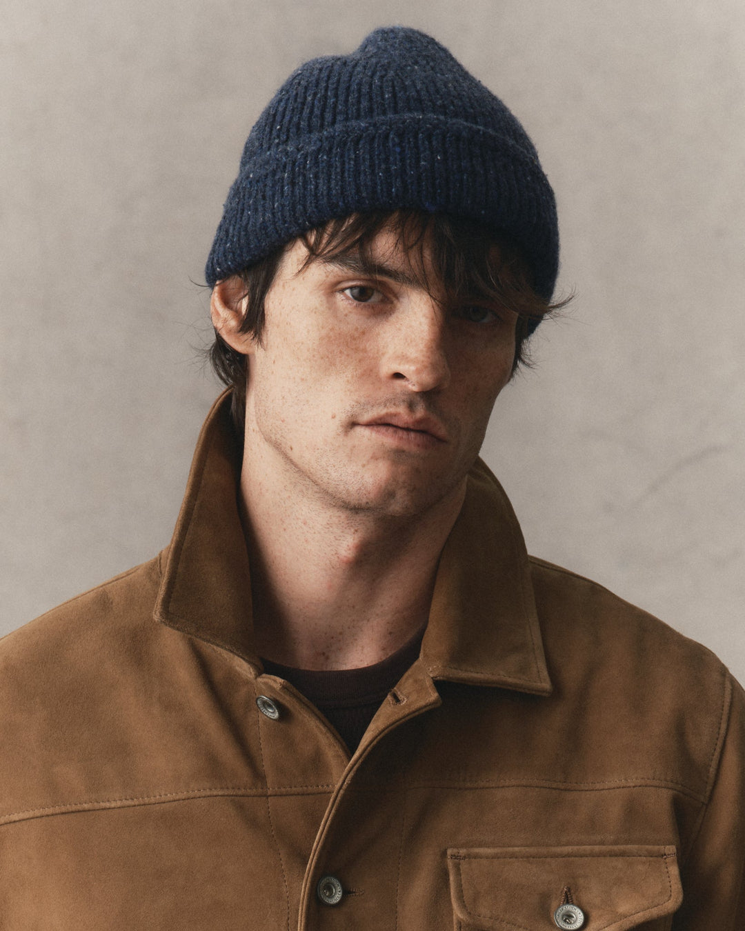 A close-up portrait of a man wearing a dark navy blue ribbed knit beanie and a brown suede jacket against a neutral textured gray background.