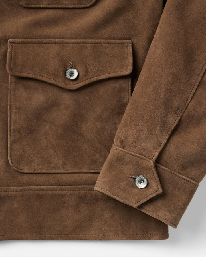 Close-up of a brown suede jacket featuring a large chest pocket with a silver snap button and a matching sleeve cuff with a snap button, shown on a light gray background.
