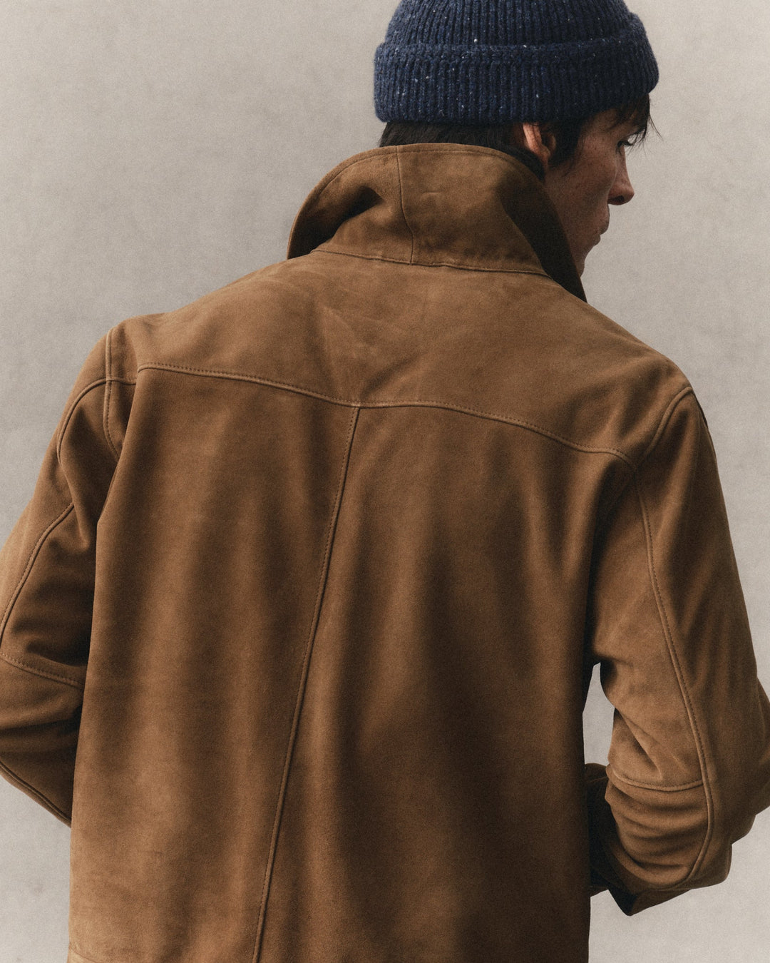 A man viewed from behind wearing a brown suede jacket and a dark navy blue knit beanie against a textured light gray wall.
