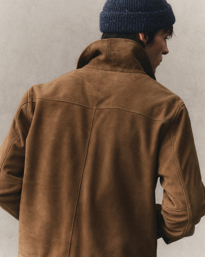 A man viewed from behind wearing a brown suede jacket and a dark navy blue knit beanie against a textured light gray wall.